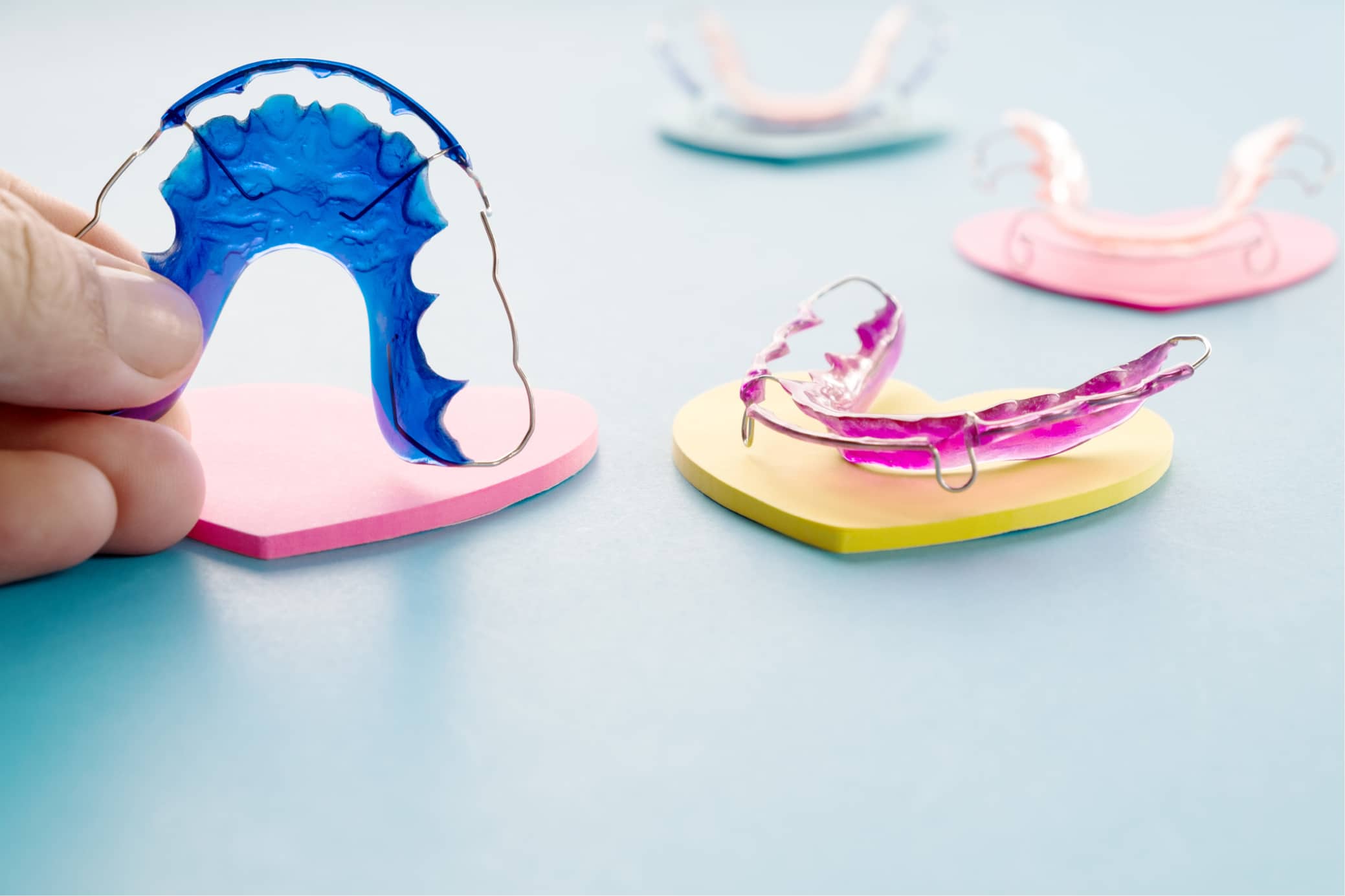 Colorful dental retainers on heart-shaped pads. A person holds a blue retainer. Background features additional retainers on a light blue surface.